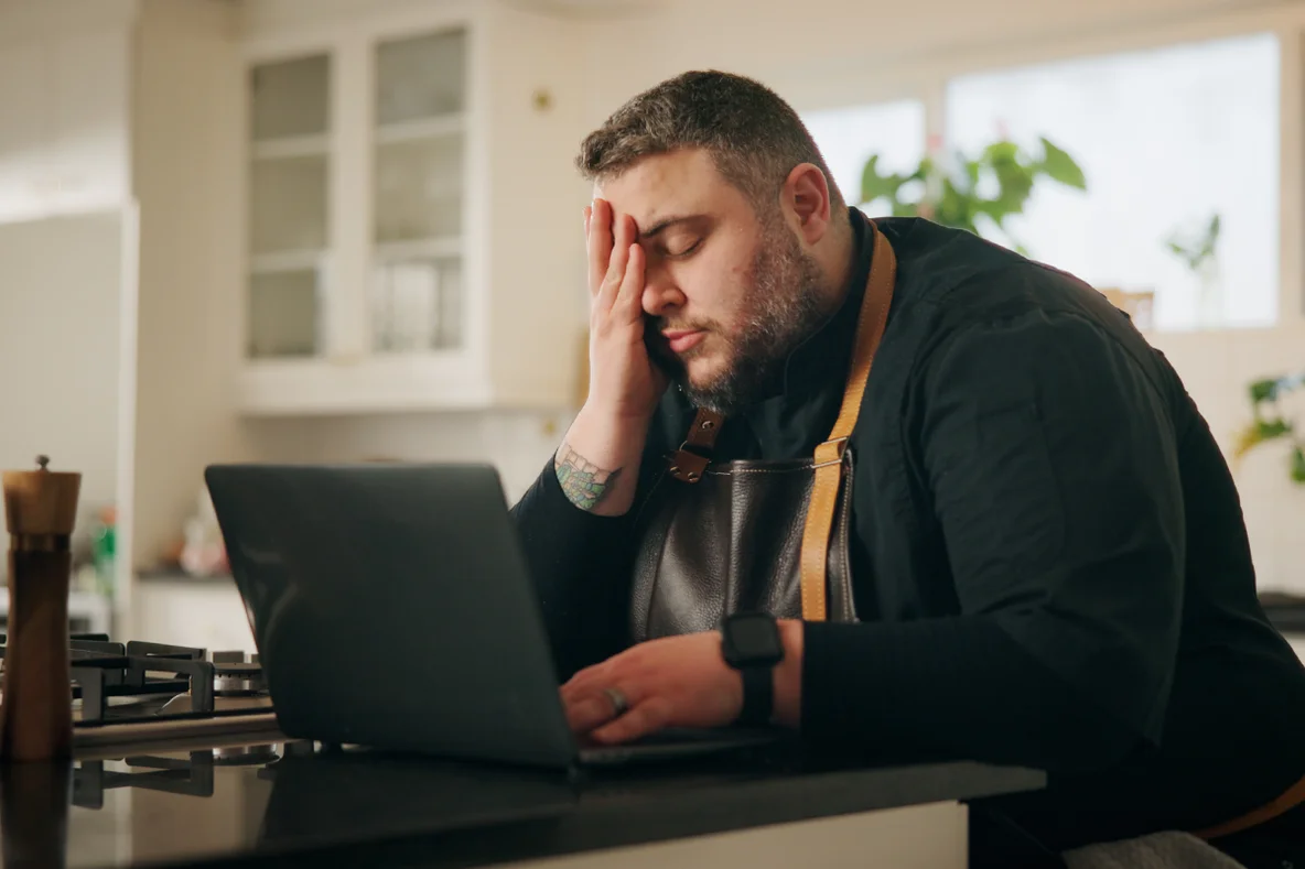Mann steht in der Küche vor einem Laptop und stützt sich müde auf die Arbeitsplatte und reibt sich die Augen.