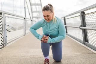 Eine Frau mit schmerzverzerrtem Gesicht kniet auf dem Boden und greift mit beiden Händen an ihr Knie. Sie trägt sportliche Kleidung.