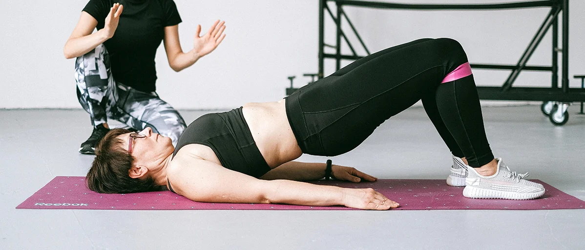 Eine Frau liegt auf einer Gymnastikmatte und hebt ihre Hüfte nach oben. Eine andere Frau sitzt daneben und gestikuliert.