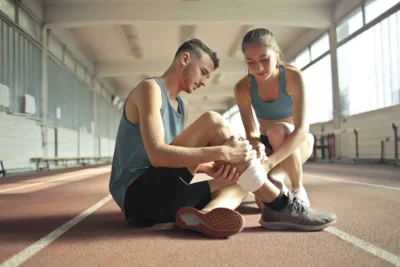 Person in Sportkleidung sitzt auf dem Boden und hält die Hand an das Schienbein. Eine andere Person hilft.