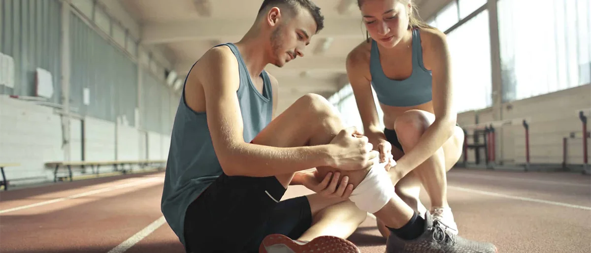 Person in Sportkleidung sitzt auf dem Boden und hält die Hand an das Schienbein. Eine andere Person hilft.