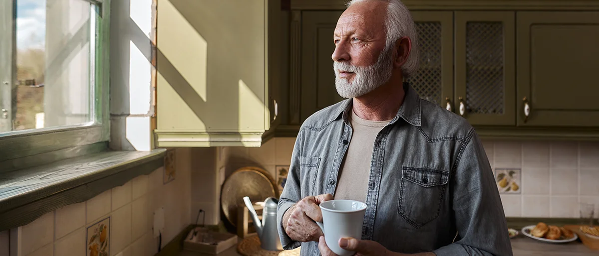 Alter Mann steht mit einer Tasse in der Hand in der Küche und schaut aus dem Fenster. Er hält die Tasse mit beiden Händen fest.