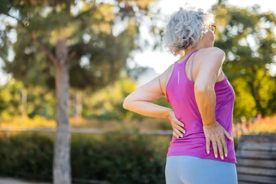Ältere, sportlich gekleidete Dame steht in einem Park und hält sich mit einer Hand an die Hüfte