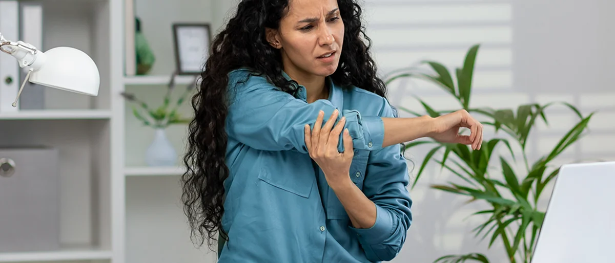 Eine Frau sitzt vor einem Laptop und greift sich mit schmerzverzerrtem Gesicht an den Ellenbogen