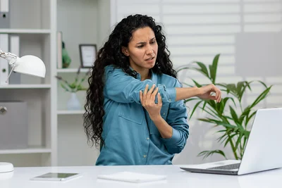 Eine Frau sitzt vor einem Laptop und greift sich mit schmerzverzerrtem Gesicht an den Ellenbogen