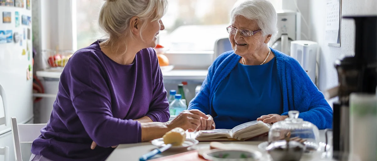 Eine alte Dame sitzt mit einer jüngeren Dame an einem Küchentisch. Sie haben vor sich ein Notizbuch liegen
