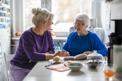 Eine alte Dame sitzt mit einer jüngeren Dame an einem Küchentisch. Sie haben vor sich ein Notizbuch liegen
