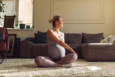 Schwangere Frau in einer eng anliegenden Leggins sitzt im Wohnzimmer auf dem Boden und dreht ihren Oberkörper.