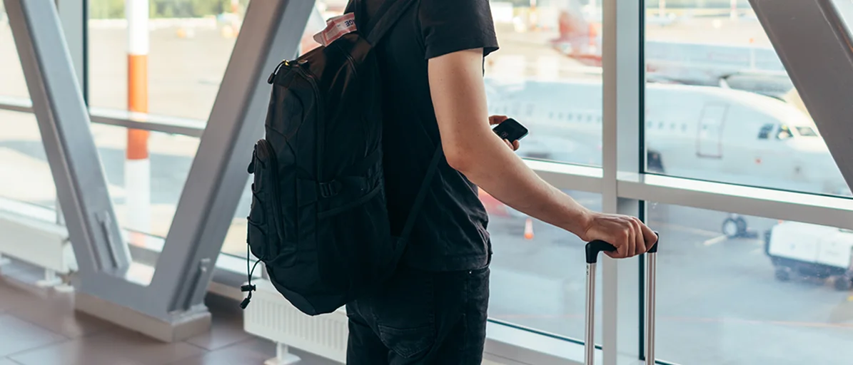 Mann steht mit Gepäck am Flughafen und schaut aus dem Fenster auf Flugzeuge