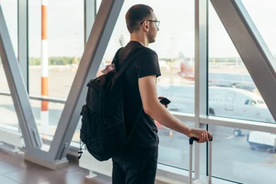 Mann steht mit Gepäck am Flughafen und schaut aus dem Fenster auf Flugzeuge