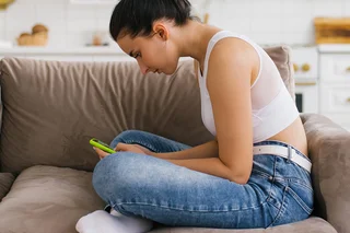 Junge Frau sitzt im Schneidersitz auf einer Couch und hat den Kopf nach unten geneigt um auf ihr Smartphone zu schauen.