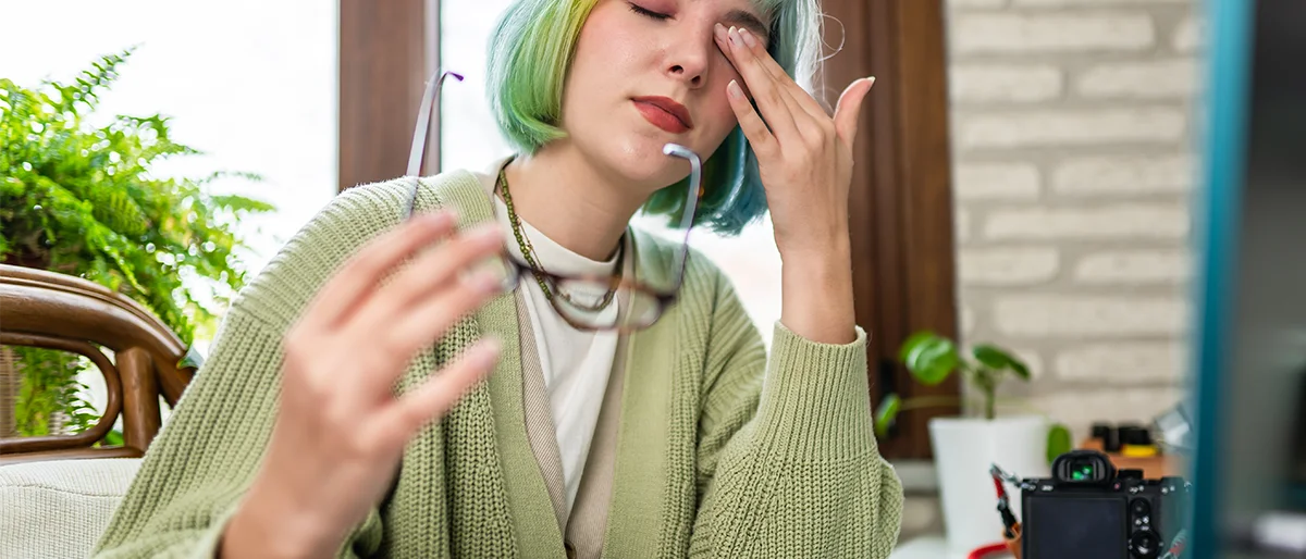 Frau sitzt am Schreibtisch und fasst sich mit einer Hand an ein Auge