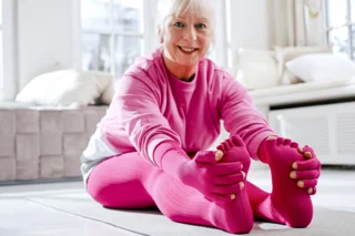 Abbildung einer älteren Dame, die sich auf dem Boden sitzend dehnt. Sie trägt pinke Kompressionsstrümpfe und -Ärmel