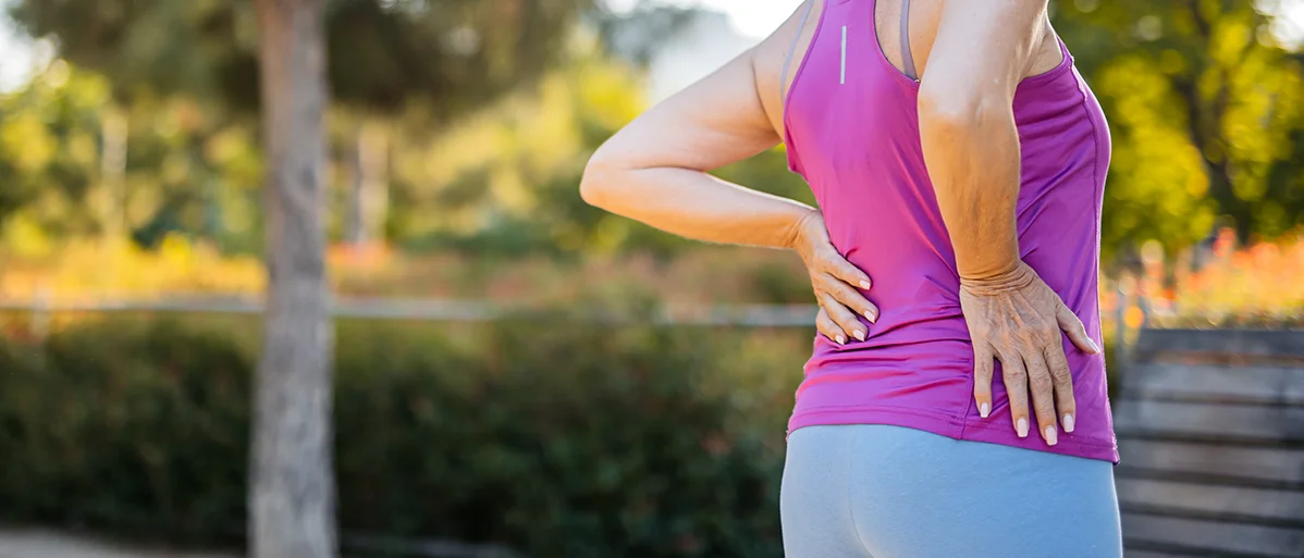Ältere, sportlich gekleidete Dame steht in einem Park und hält sich mit einer Hand an die Hüfte