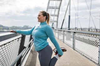 Eine Frau in sportlicher Kleidung steht an dem Geländer einer Brücke. Sie zieht ihren Fuß mit der Hand Richtung Gesäß um ihren Oberschenkel zu dehnen.