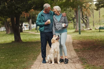 Zwei ältere Personen stehen auf einem Gehweg und halten sich lachend in den Armen. Sie haben beide einen Kaffeebecher in der Hand.