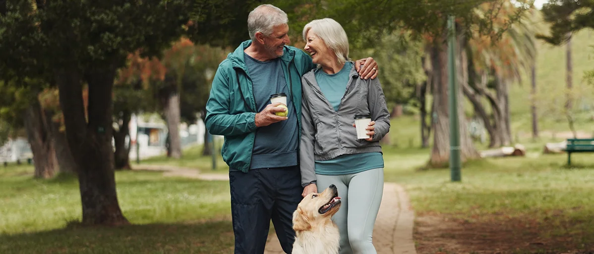 Zwei ältere Personen stehen auf einem Gehweg und halten sich lachend in den Armen. Sie haben beide einen Kaffeebecher in der Hand.