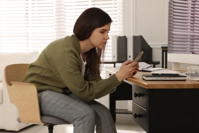 Eine Frau sitzt mit gekrümmten Rücken auf einem Stuhl. Sie hat ein Smartphone in der Hand.