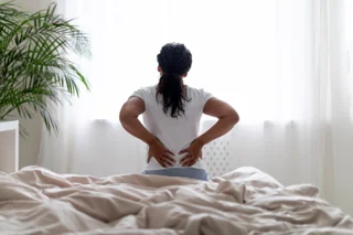 Person sitzt auf dem Bett mit dem Rücken zum Betrachter und greift sich mit beiden Händen an den Rücken.