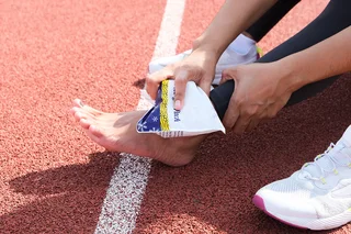 Eine Person sitzt auf dem Boden eines Sportplatzes und kühlt sich den Knöchel am Fuß