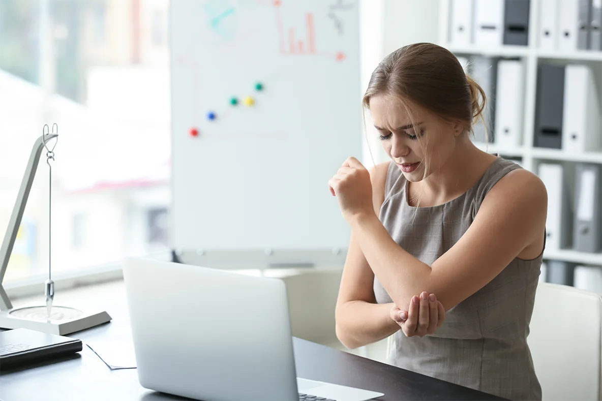 Abbildung einer Frau, die vor einem Laptop am Schreibtisch sitzt und sich an den Ellenbogen greift