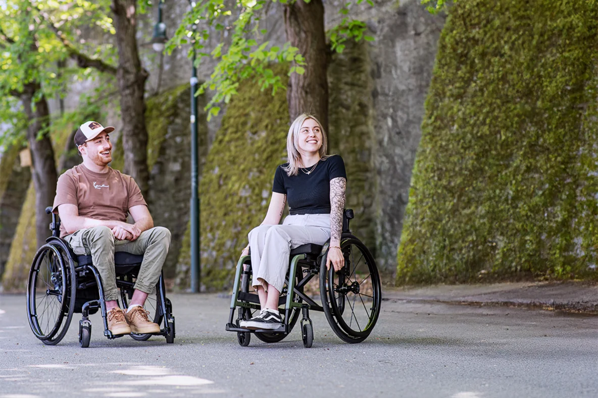 Ein Mann und eine Frau sitzen im Rollstuhl und sind draußen unterwegs