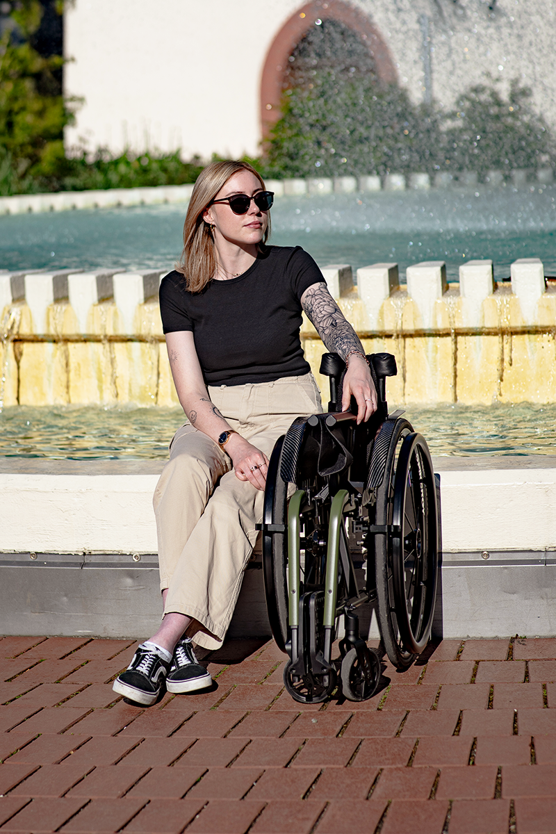 Frau sitzt an einem Brunnen. Neben ihr steht ihr zusammengeklappter Rollstuhl.